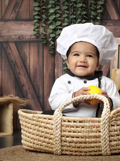 A smile that melts hearts. This little chef is happily sitting in a basket, ready to get cooking in a rustic kitchen setup designed for a six-month or one-year-old baby photoshoot.