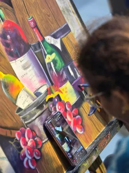 A student uses her phone as a reference for her still life painting. This piece, featuring wine bottles and grapes, is a great exercise in capturing color, light, and reflection.