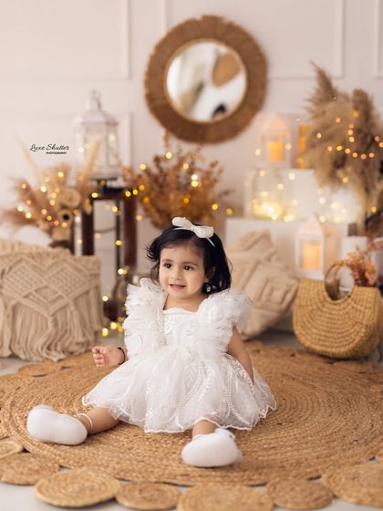 A sweet, smiling portrait of the birthday girl in a white lace dress, sitting on a jute rug in a cozy, bohemian-themed studio setup.
