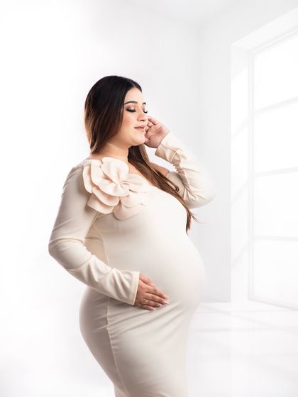 A serene close-up of a mother-to-be in a fitted cream gown. The soft, natural light from the window highlights her gentle expression and the beautiful floral detail on the dress.