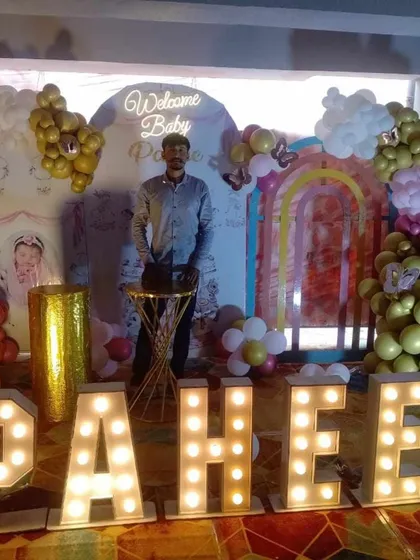 A grand 'Welcome Baby' setup featuring large marquee letters that spell out the baby's name, 'PAHEE'. The backdrop includes a neon sign, a rainbow arch, and balloon clouds for a spectacular welcome.