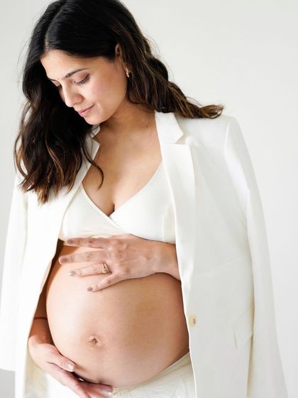A close-up portrait of a pregnant mother in a stylish white blazer, looking down at her bump.