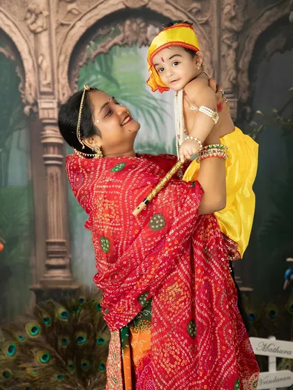 A mother dressed in a vibrant red bandhani saree joyfully lifts her baby, who is dressed as little Krishna, during a festive Janmashtami photoshoot.