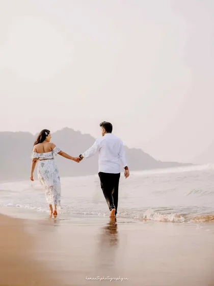 A couple walks away from the camera along the wet sand, holding hands as they look out at the ocean and mountains in the distance.