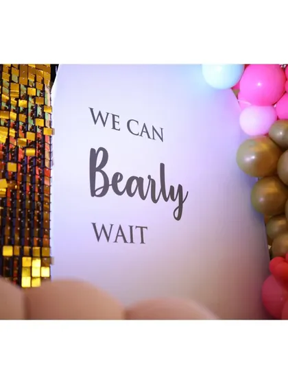 A close-up of the baby shower backdrop, clearly showing the theme "We Can Bearly Wait" next to a shimmering gold sequin panel.