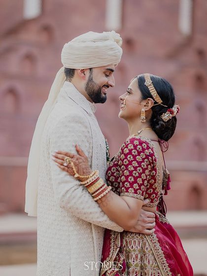 Against the historic backdrop of a red stone fort, this couple shares a look of pure love. Their classic wedding attire, a white sherwani and a red lehenga, creates a striking and timeless portrait.
