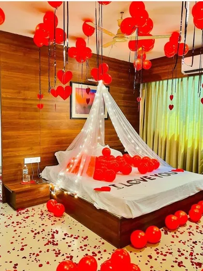 I create deeply romantic moments with setups like this. The room is filled with red balloons, a floor scattered with rose petals, and a bed featuring "I LOVE YOU" spelled out, all under a dreamy, lit canopy.