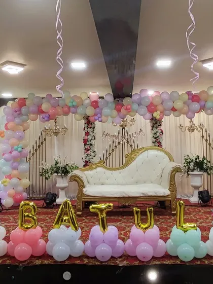 An elegant and grand first birthday setup in a party hall. This design includes a wide pastel-colored organic balloon arch over a regal white chaise lounge, with the name "BATUL" spelled out in balloon letters at the front.