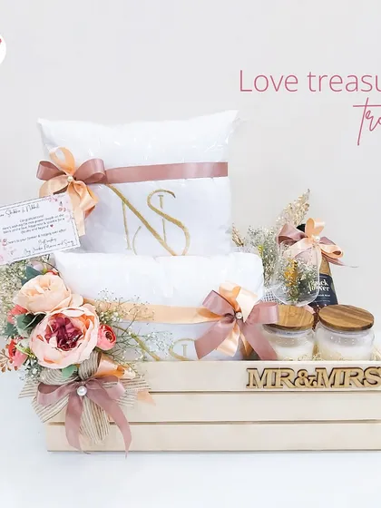 A "Love Treasure Trove" hamper for the happy couple. This crate includes personalized "Mr & Mrs" items, monogrammed pillows and towels, and other curated goodies, adorned with soft peach florals.