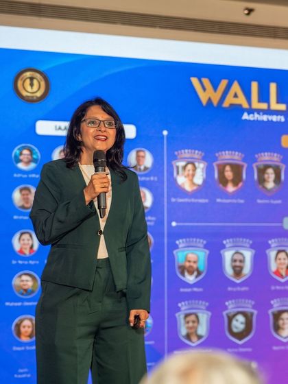 Speaking on stage in a green suit in front of the 'Wall of Achievers'. Degrees may open doors, but true growth comes from continuous learning beyond the classroom.