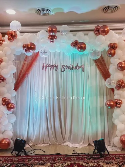 An elegant backdrop for an 89th birthday celebration. This setup features a full balloon arch in white and rose gold chrome, with a simple 'Happy Birthday' foil banner and dramatic uplighting against a curtained backdrop.