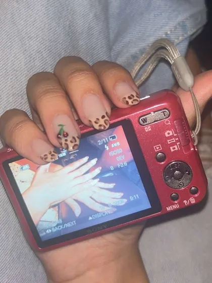 A fun and trendy set featuring leopard print nails with a cherry art accent, perfectly posed with a retro digital camera for that Y2K aesthetic.