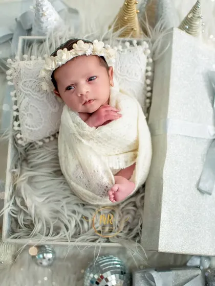A beautifully wrapped newborn lies in a gift box, surrounded by sparkling silver disco balls and Christmas decorations.