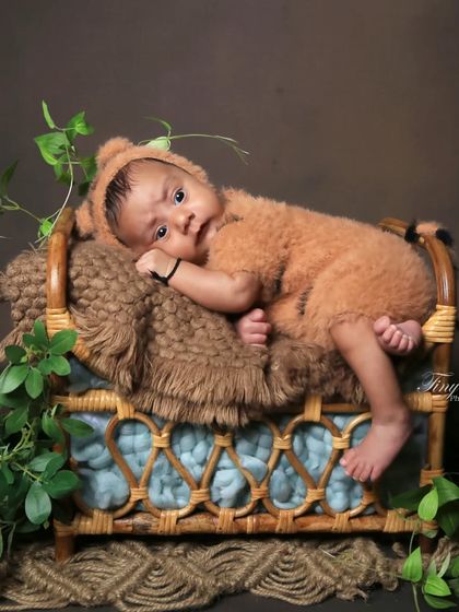 This "bear cub" photo shows an awake baby in a fuzzy brown outfit, resting on a tiny cane bed. The setup is styled with natural textures and green vines for a rustic, woodsy feel.