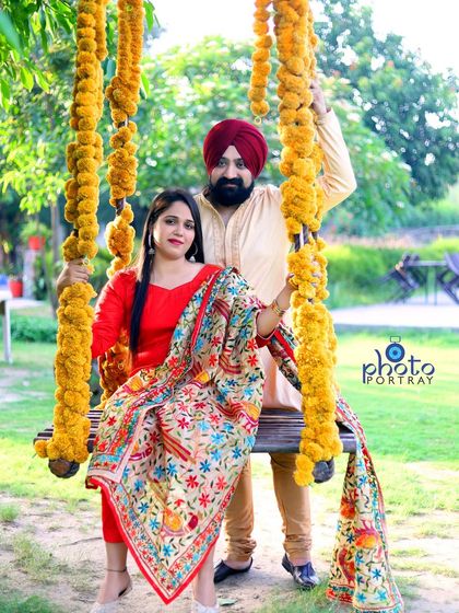 A vibrant and traditional pre-wedding photo. The couple is dressed in colorful ethnic wear, posing on a swing decorated with marigold flowers for a festive feel.