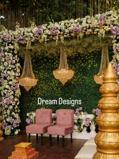 A close-up of the mandap seating, featuring plush pink chairs and traditional golden pillars. The backdrop is a wall of greenery illuminated by tiny lights, with three crystal chandeliers adding a touch of opulence.