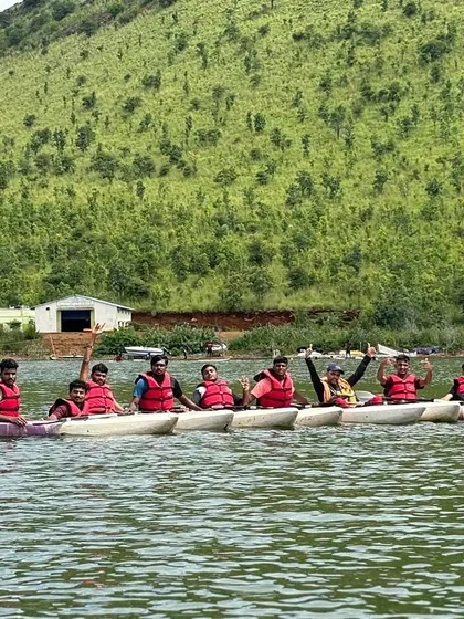 A team of kayakers waves from the water, with the green hills of Vani Vilas Sagar providing a beautiful backdrop.
