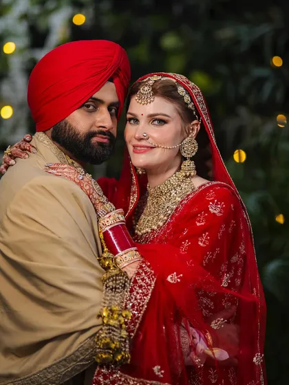 This portrait from a beautiful NRI wedding showcases the couple's regal attire and their comfortable connection. The bride's traditional red lehenga and the groom's turban create a stunning visual against the soft, natural background.