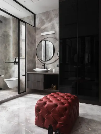 This bathroom combines moody grey marble with sleek, black-tinted glass for the wardrobe and shower partition. A pop of color from the red leather ottoman adds an unexpected and luxurious touch.