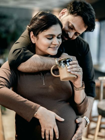 A cozy and intimate indoor moment. He embraces her from behind as she holds a warm mug, both of them smiling and content.