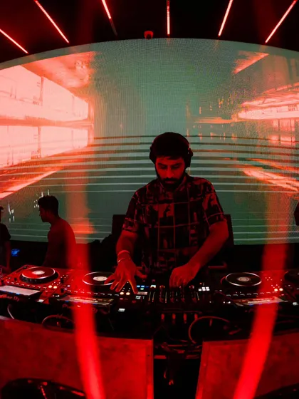 Focused on the mix, with the red lights of the club creating a dramatic backdrop. Every track is carefully selected to build the energy and keep the journey going.
