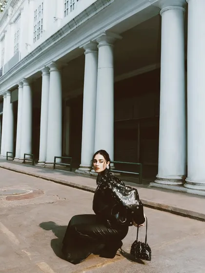 A creative, low-angle shot of Kavya Trehan. The grand pillars create a dramatic backdrop for this edgy, all-black fashion look.