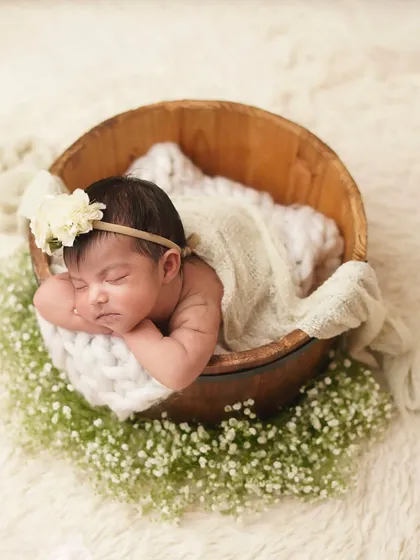A newborn girl sleeps in a rustic wooden bucket, surrounded by a bed of baby's breath. This setup combines natural elements to create a beautiful, garden-themed infant portrait.