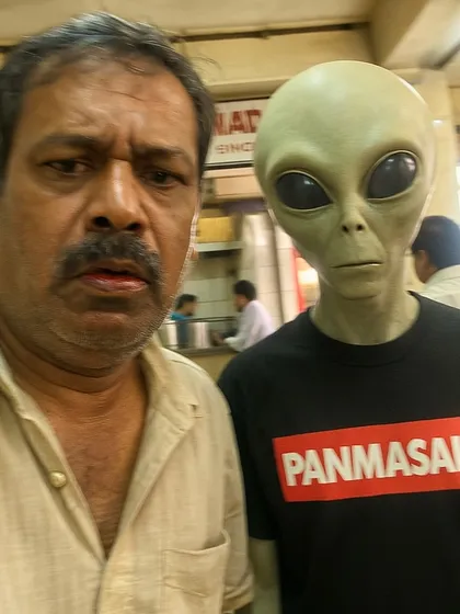 A man takes a disgruntled selfie with an alien wearing a 'PANMASALA' t-shirt. A classic 'Riyal Life' image, finding humor in the mundane collision of the otherworldly and the everyday.