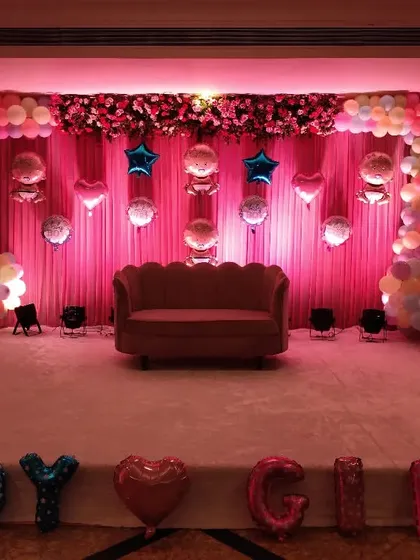 An elegant stage setup for a baby shower, using soft pink lighting and drapes to create a warm ambiance. The decoration includes a pastel balloon arch, a floral border, and hanging foil balloons, with large 'BOY' or 'GIRL' foil letters in front, making it perfect for a gender reveal party.