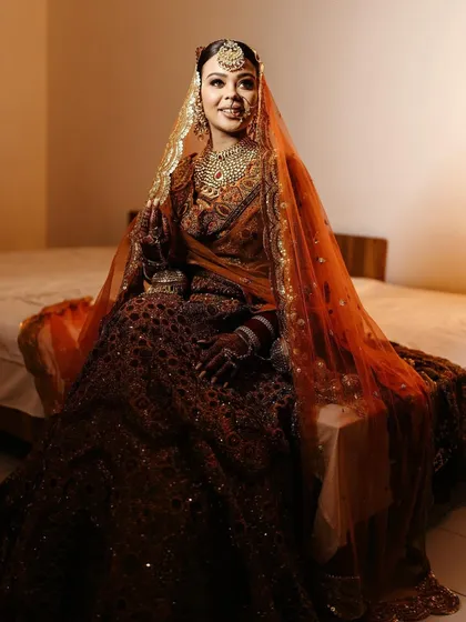 A full-length portrait of the bride seated on a bed, her rich, dark lehenga and orange dupatta creating a stunning, regal look.