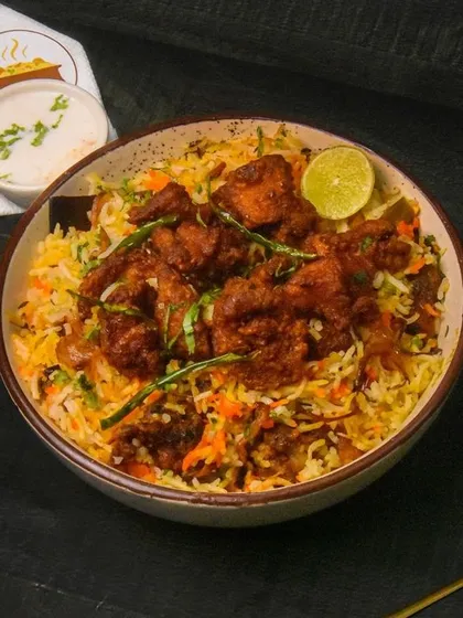 A bowl of spicy chicken biryani, garnished with a lime wedge and served with raita. The dark background makes this a rich and inviting photograph for an Indian restaurant.