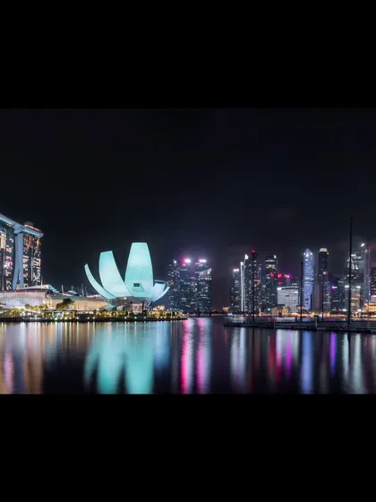 A panoramic night photograph of Singapore's Marina Bay, capturing the iconic skyline and its reflection in the water.