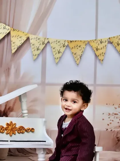 A little musician in the making, this toddler smiles as he sits by a miniature white piano in a beautifully decorated room.