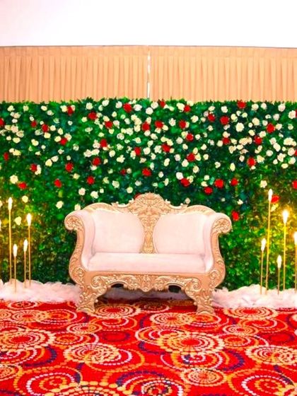 A simple and elegant wedding reception stage. The setup features a green leaf wall with red and white roses, fronted by a classic white sofa.