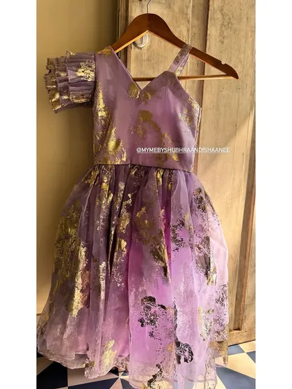 A magical party dress for a little princess. This lavender creation features a gold foil print on a sheer overlay, an asymmetrical neckline, and a single ruffled sleeve for a touch of whimsy.