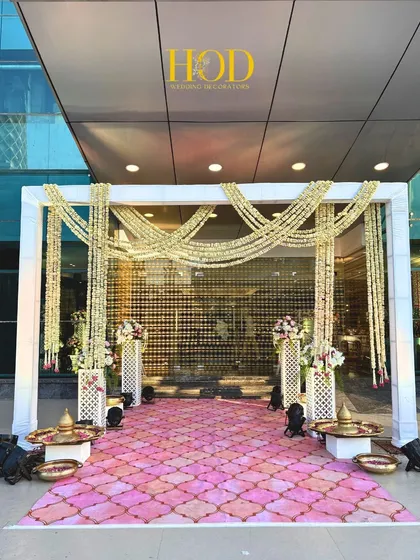 A beautiful outdoor entrance setup with a white frame, draped with jasmine flower strings, leading to a vibrant pink patterned floor for a traditional yet fresh look.