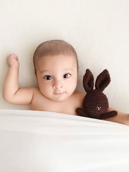 A baby lies on a white blanket with a small knitted bunny prop.