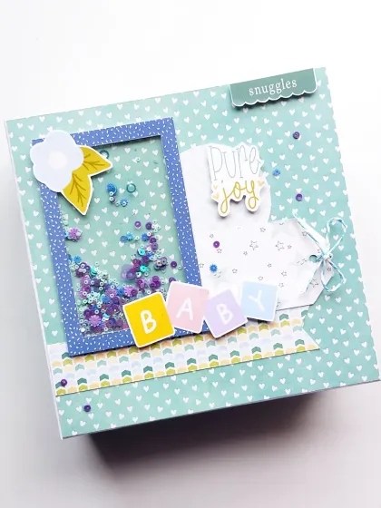 Celebrating a new arrival is pure joy. This is the cover of a baby scrapbook, designed with a fun shaker window filled with colorful sequins and cute "BABY" blocks to mark the beginning of a beautiful new story.