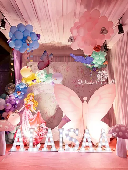 A beautiful photo booth for a princess-themed party, featuring a large illuminated butterfly, marquee letters spelling "ALAISAA," and a cutout of Princess Aurora. The pink and pastel color scheme creates a dreamy atmosphere.