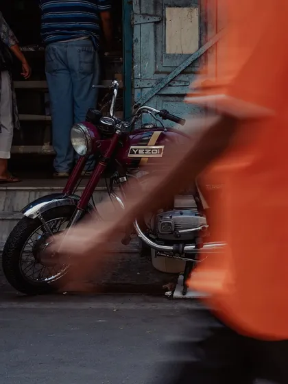 A vintage maroon Yezdi motorcycle is captured in sharp focus while the motion of a person walking past is blurred into an orange streak. This artistic technique freezes a moment in time, contrasting the timeless bike with the city's constant movement.