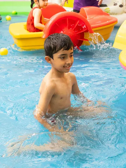 A young boy enjoys the cool water of the splash pool, with a colorful paddle boat visible in the background. There are so many different ways to play at our summer events.