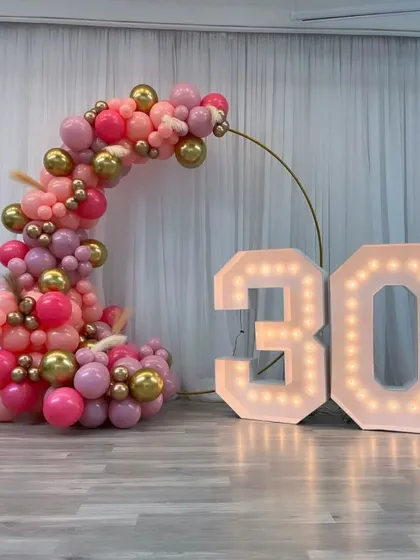 A beautiful crescent-shaped balloon arrangement for a 30th birthday. The pink and gold balloons are paired with large light-up marquee numbers for a stylish and modern look.