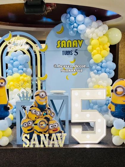 A fun-filled Minions theme for a 5th birthday party. The setup includes multiple Minion character cutouts, a large light-up number '5', and a blue and yellow balloon arch decorated with bananas.