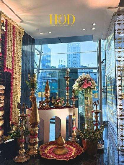 A detailed view of a traditional decor corner at an entrance, featuring brass diyas, floral urlis, and hanging flower strings against a modern glass window.