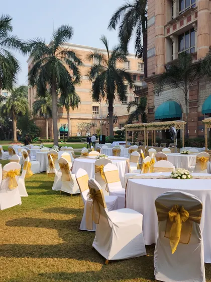 Guest seating for a sunny outdoor luncheon. The round tables are covered in white linen, and the chairs are decorated with golden sashes, creating a cheerful and elegant look.
