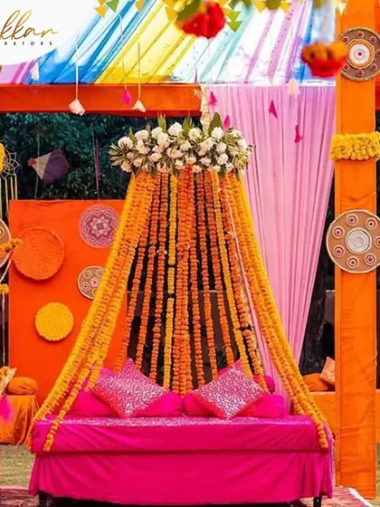 A vibrant Mehendi seating arrangement with a canopy of marigold strings. The bright pink sofa contrasts beautifully with the orange decor, creating a lively and colorful space.