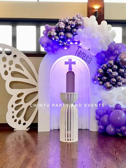 A butterfly-themed First Holy Communion decoration in shades of purple. The setup includes a large butterfly wing cutout and a lighted arch backdrop.