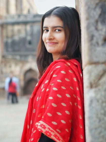 A warm and friendly portrait leaning against a historic pillar. Her smile and the classic black suit with a red dupatta create a timeless and approachable look.