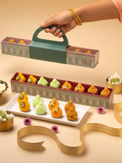 This photo showcases an elegant, modern gift box for modaks. I styled it with a golden ribbon and fresh flowers on a neutral background to highlight the unique packaging and the beautiful arrangement of the sweets.
