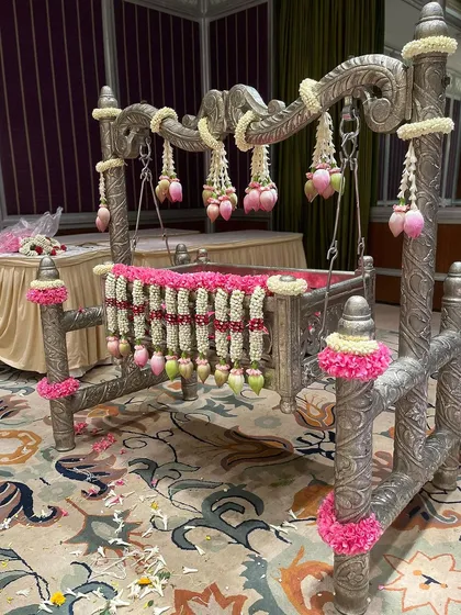A silver cradle decorated with jasmine, pink flowers, and lotus bud garlands, with scattered petals on the floor after a beautiful naming ceremony.